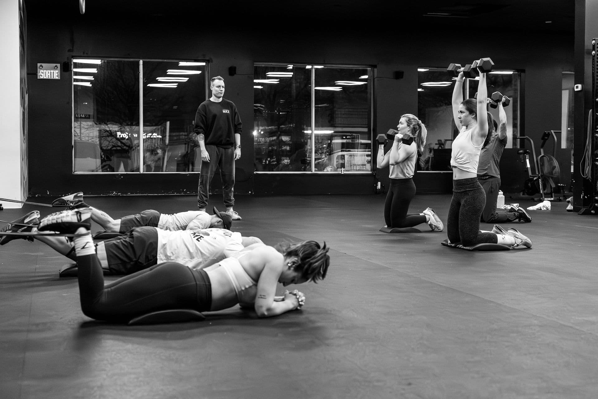 A class doing kneeling dumbbell presses, with a coach at the back.